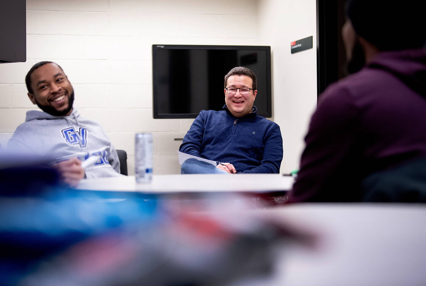 Three individuals are seated around a table in a meeting room, smiling and engaged in conversation.